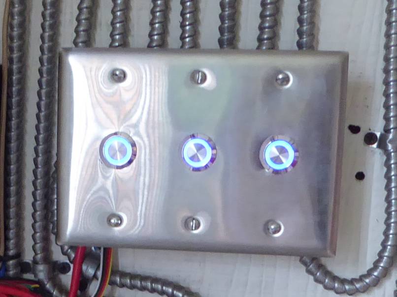 Image of three buttons with blue backlight mounted onto stainless steel plate in a row, surface mount device box